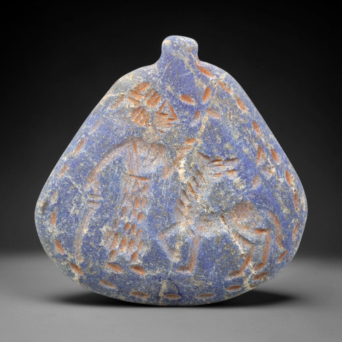 Ancient Lapis Amulet - Near Eastern Figural Scene, 79ZY: Carved from lapis lazuli, the amulet features an incised figural scene depicting a humanoid figure alongside a quadrupedal animal, rendered in raised relief with reddish mineral inlay that accentuates