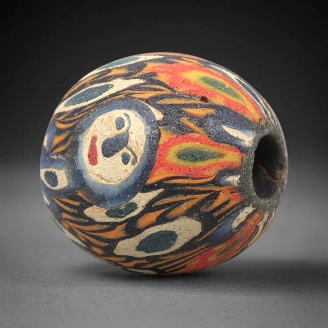 Ancient Mosaic Glass Bead - Roman Face Pattern Amulet, 58WD: This spherical bead is crafted from core-formed mosaic glass, featuring a vibrant multi-color base decorated with intricate figural face motifs and swirling foliate elements. Each face motif consists