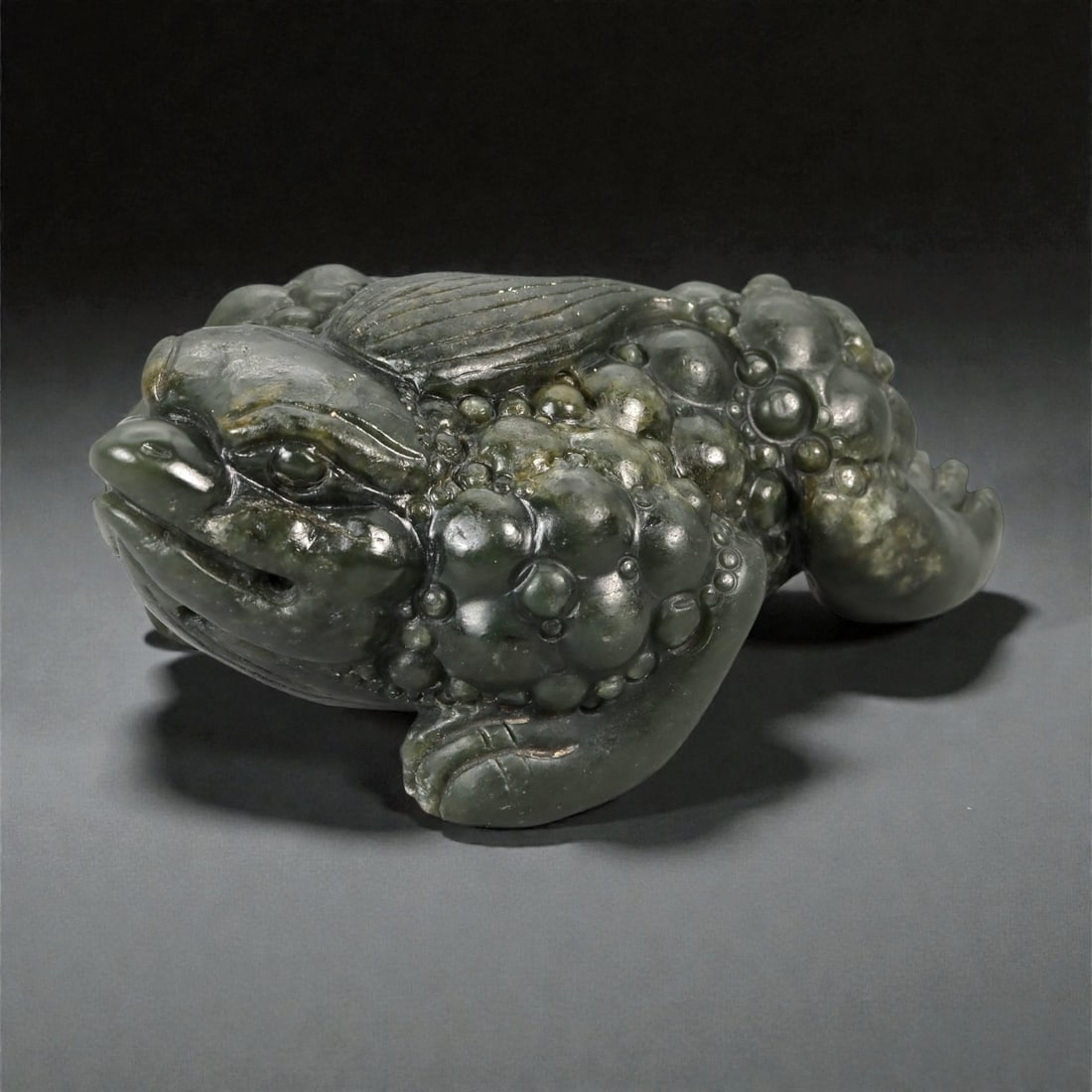 A Chinese Nephrite Jade Frog Carving, 20th Century, 25FB: Carved from dark green nephrite jade with lighter mottling and natural inclusions, the sculpture is worked in the round in the form of a crouching frog. The body is compact and low to the ground, with