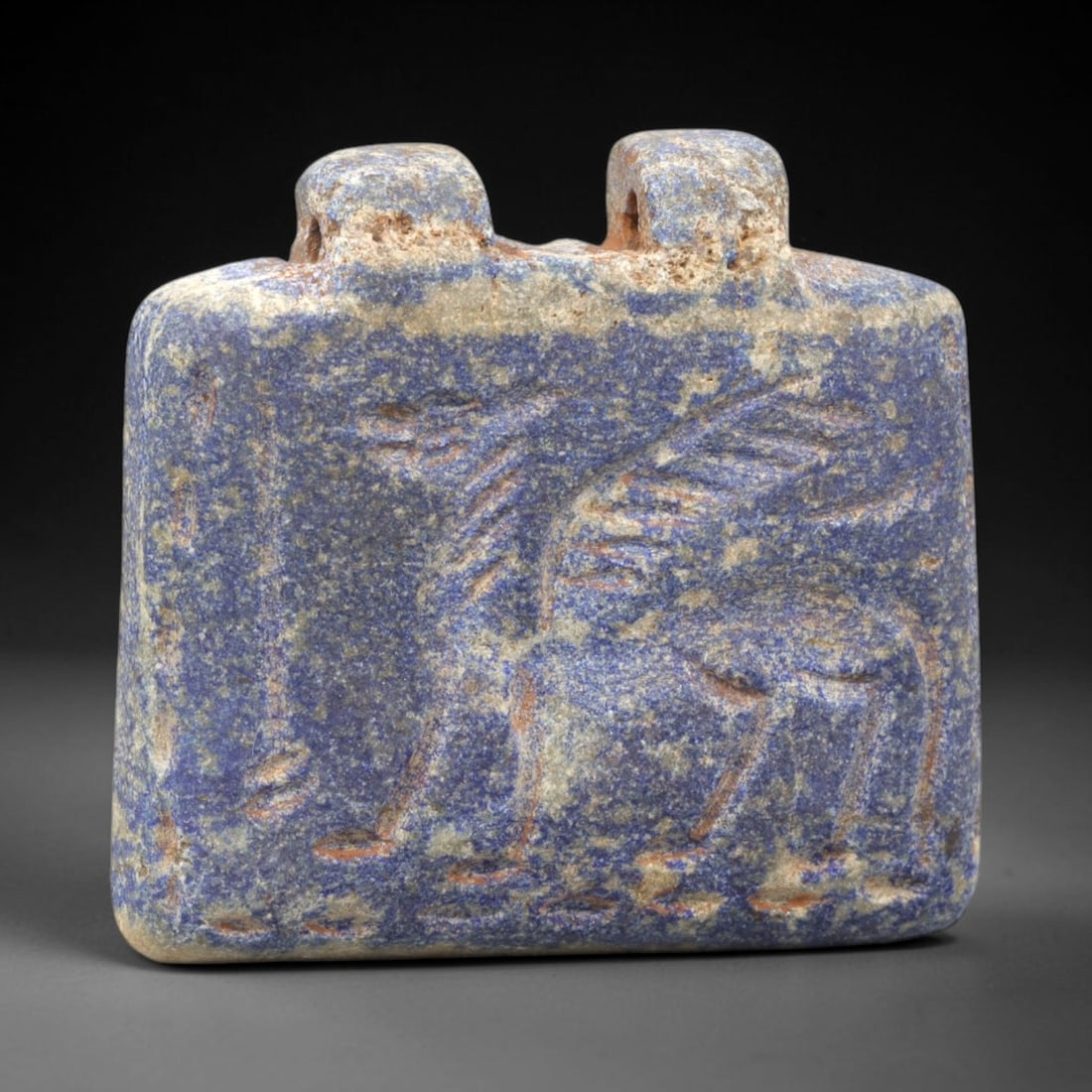Ancient Lapis Amulet - Near Eastern Animal Figure, 82ZY: Carved from lapis lazuli, the amulet is shaped as a flat rectangular plaque with two perforated suspension knobs on the upper edge, designed for wear. The front face is incised in low relief with a st