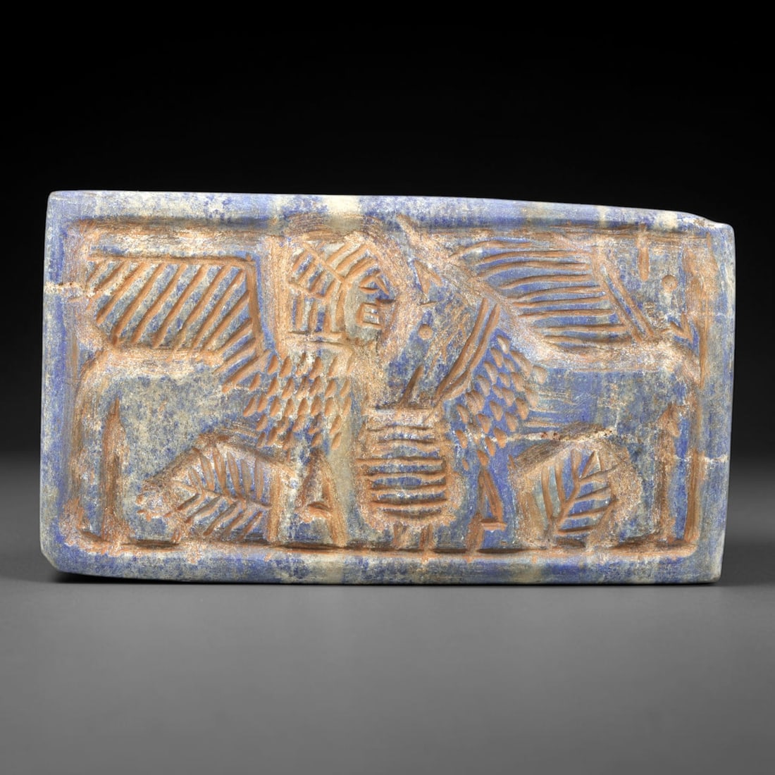 Near Eastern Lapis Lazuli Plaque - Tribal Art Figure & Two Animal Relief, 39ZA: Carved from lapis lazuli, this rectangular plaque features an 8.3x4.6cm (3.27x1.81in) relief composition centered on a stylized human figure, flanked by two quadrupedal animal motifs, executed in the