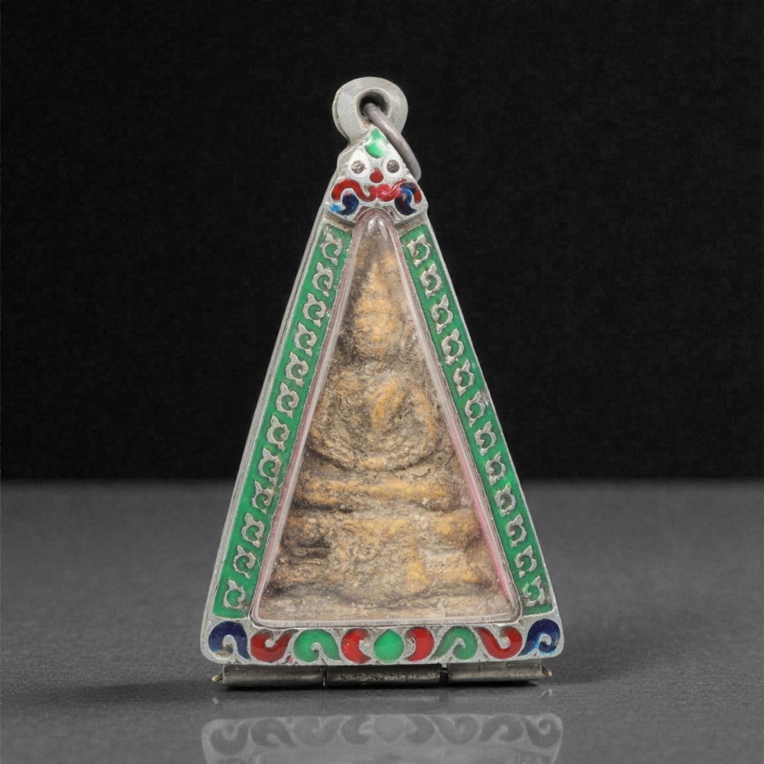 A Thai Buddha Amulet Pendant, 20th Century, 2FP: This triangular pendant encloses a seated Buddha amulet core within a silver frame. The obverse frame is adorned with vibrant green, red, and blue enamel scrollwork, while the reverse frame displays i