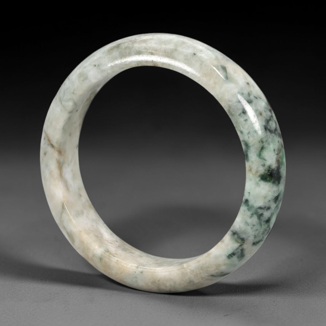 A Chinese Jadeite Jade Bangle Bracelet, 20th Century, 84ZK: Carved from jadeite jade with a pale off-white base and irregular dark green mottled inclusions, the bangle is shaped as a solid, continuous circular band with a classic rounded cross-section. The sur