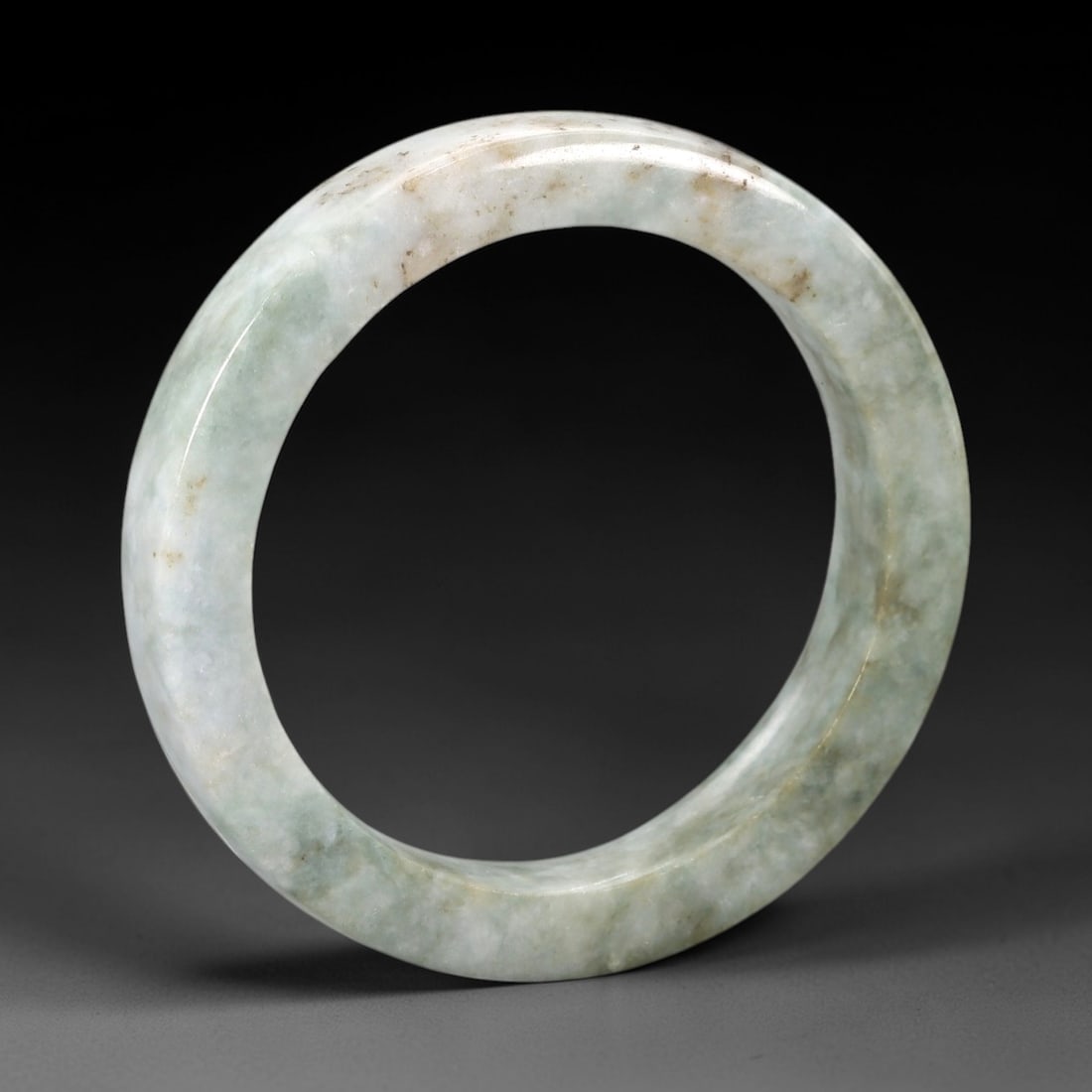 A Chinese Jadeite Jade Bangle Bracelet, 20th Century, 85ZK: Carved from jadeite jade with a pale greenish-white base and scattered light brownish inclusions, the bangle is formed as a continuous circular band with a rounded cross-section. The surface is smooth