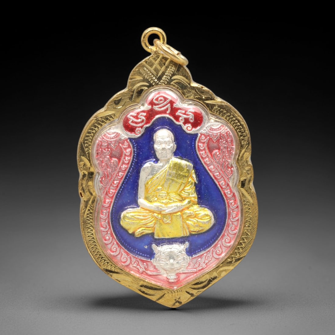 A Thai Monk Commemorative Amulet in a Gilded Case, Late 20th Century, 21ZK: This ornate amulet is presented in a gilded metal case with a suspension loop at the apex. The obverse features a vibrant, multi-colored relief of a seated Thai Buddhist monk, clad in golden robes, se