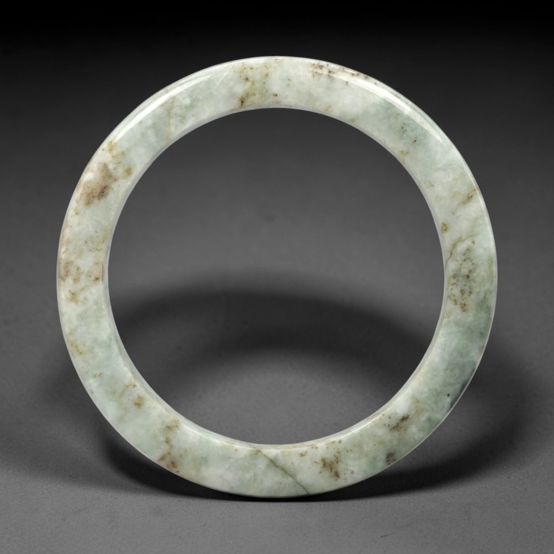 A Chinese Jadeite Jade Bangle Bracelet, 20th Century, 1ZL: Carved from jadeite jade with a pale green base and scattered brownish inclusions, the bangle is formed as a continuous circular band with a rounded cross-section. The surface is smoothly polished, wi
