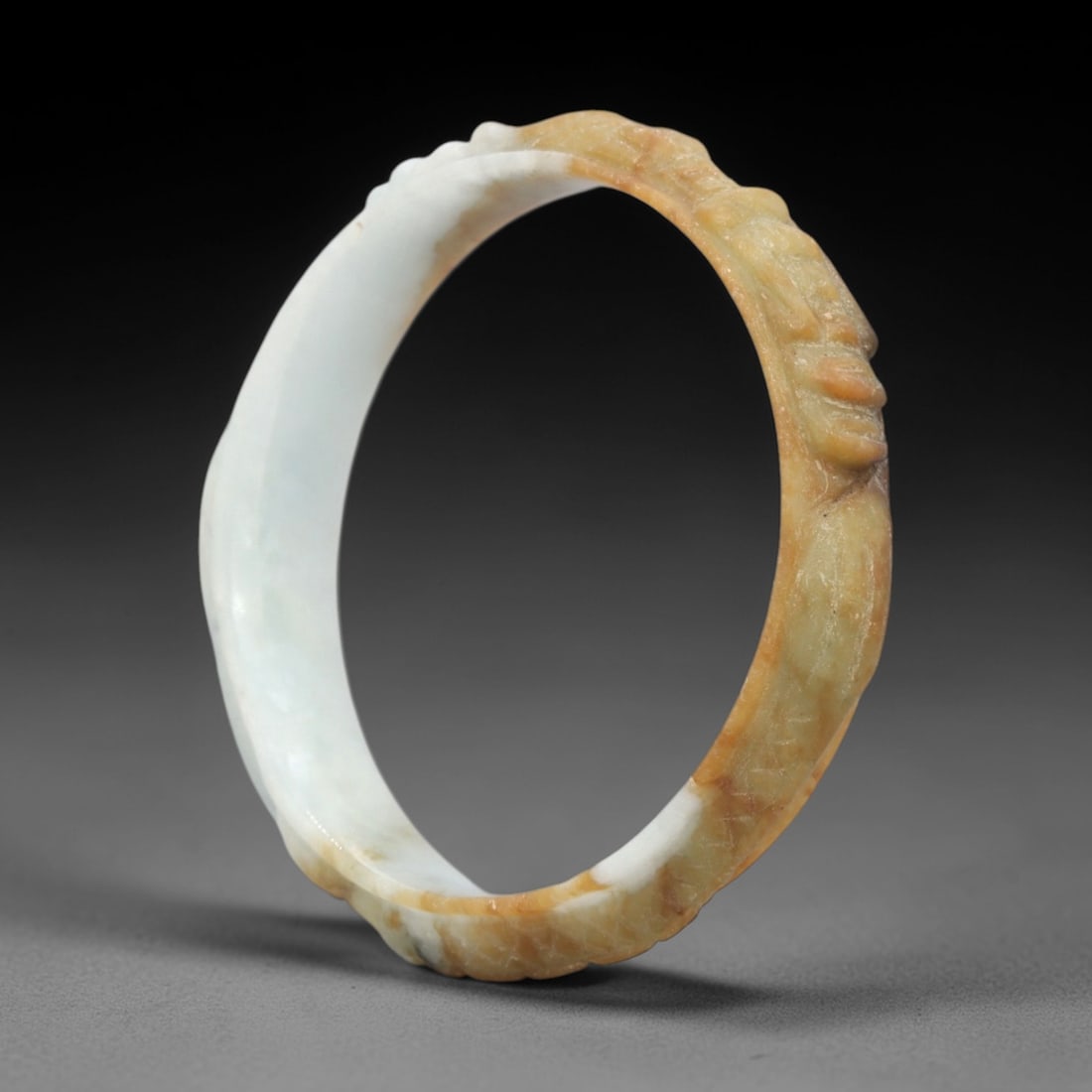 A Chinese Jadeite Jade Bangle Bracelet, 20th Century, 33FS: Carved from variegated jadeite jade with natural pale white and golden-yellow tonal variation, the bangle is formed as a continuous circular band. The exterior surface is decorated with raised relief