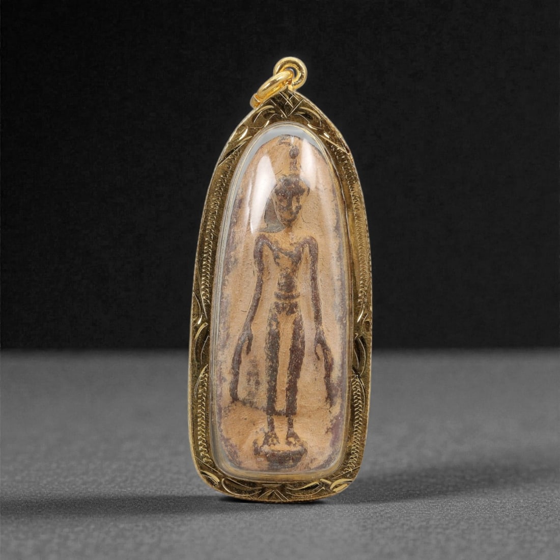 A Thai Standing Buddha Amulet with Gilt-Metal Frame, 20th Century, 33FP: The amulet is of elongated, teardrop-shaped form, featuring a molded standing Buddha figure on the obverse, rendered in a dark patinated material against a light beige ground. The Buddha is depicted w