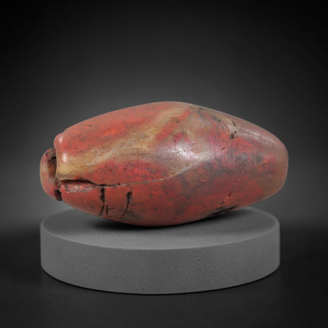 Ancient Jasper Bead - Western Asiatic Carving, 55BC: This artifact is crafted from natural jasper, displaying rich, saturated red and deep ochre mineral hues with subtle cream and grey tonal veining throughout the polished surface. The bead is formed in