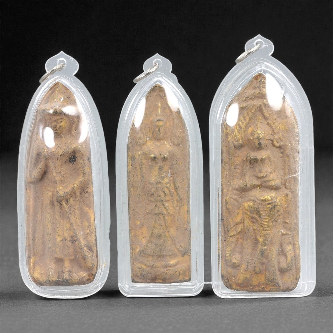 A Group of Three Gilt Buddhist Amulets in Acrylic Cases, 20th Century, 21FJ: The group comprises three elongated Buddhist amulets, each cast or molded in a gilt metal or alloy and enclosed within clear molded acrylic pendant cases fitted with suspension rings. The amulets are