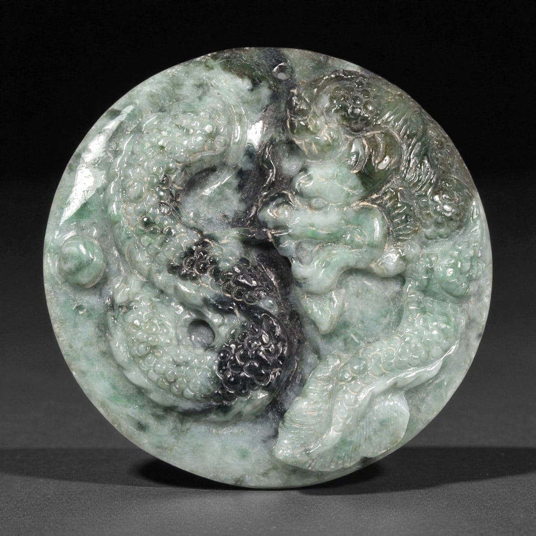 A Chinese Jade Carved Roundel, 20th Century, 4ZB: Carved from mottled green and dark-toned jade, the roundel is worked in high relief with a sinuous dragon shown in profile encircling a central aperture. The surface displays graduated scaling, a ridg