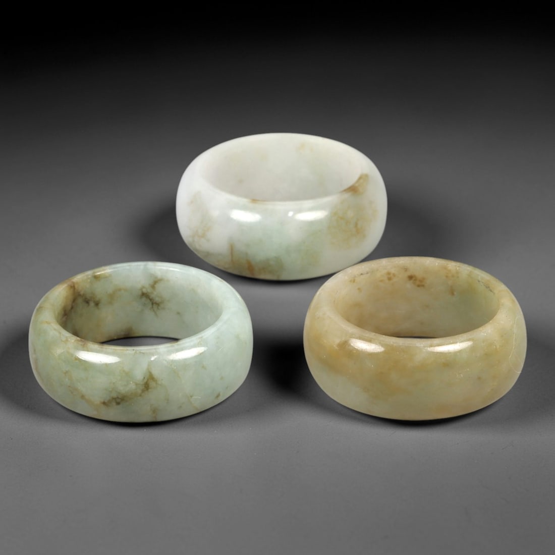 A Trio of Chinese Jadeite Rings - 20th Century, 29AL: This set of three Chinese jadeite rings showcases thick, rounded band forms carved from natural jadeite in pale celadon, milky white, and light amber tonalities, each accented by delicate mineral vein
