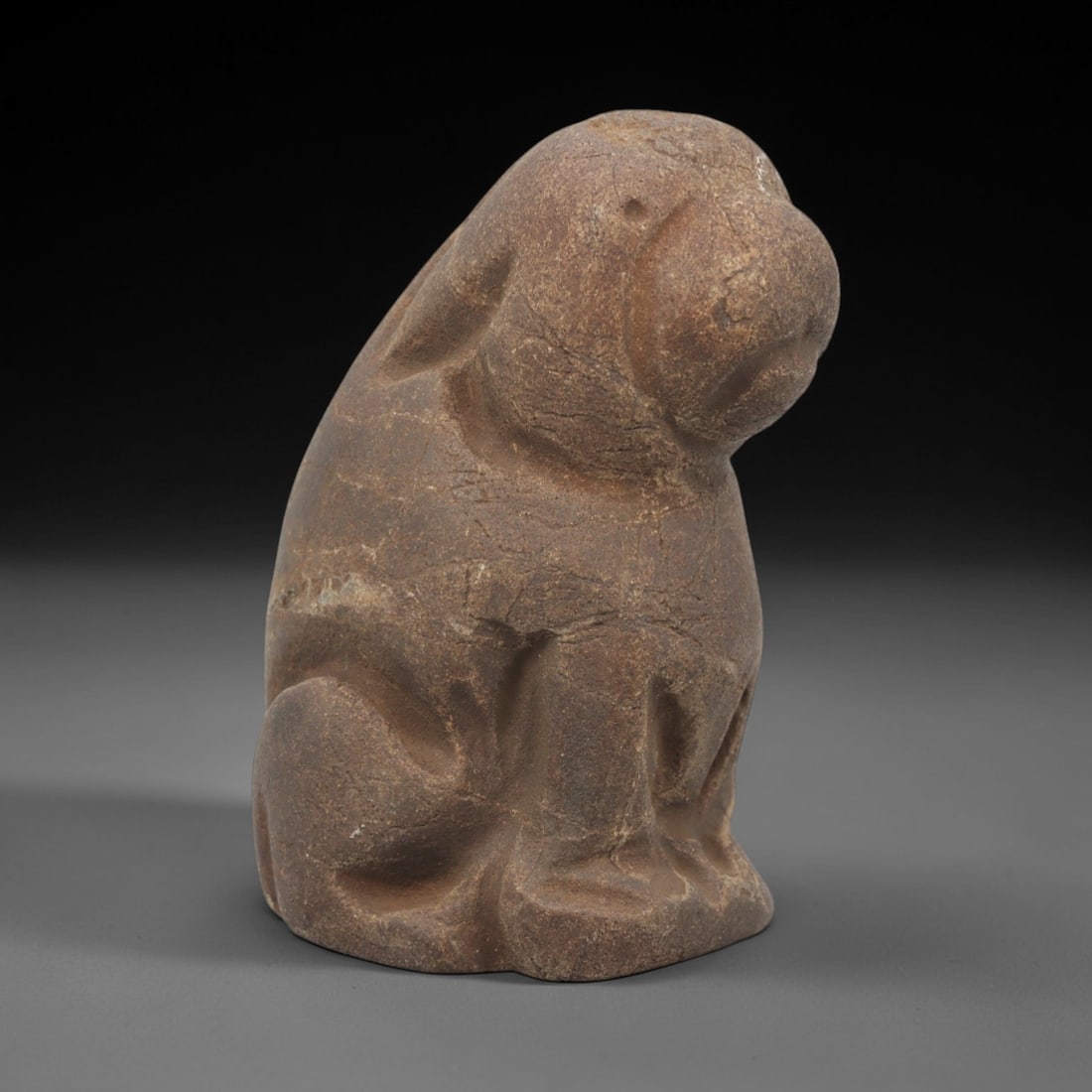 Ancient Stone Canine - Near Eastern Animal Figurine, 51FX: Carved from a coarse, earthy stone, this seated canine figure exhibits simplified, rounded forms with incised lines defining the limbs, facial contours, and ears. The compact, robust posture and styli