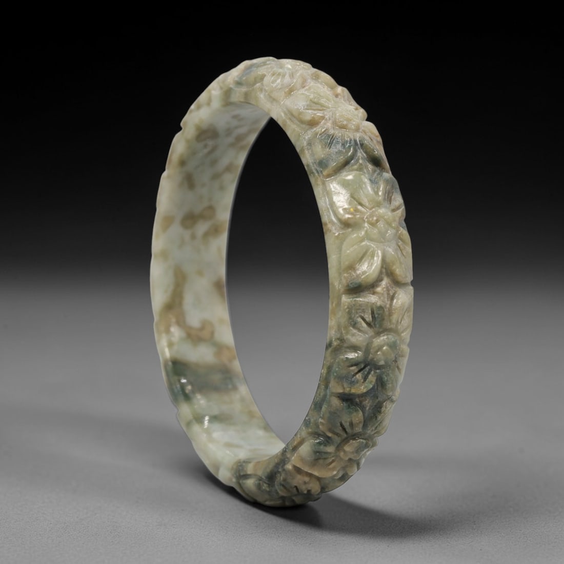 A Chinese Carved Jadeite Jade Bangle Bracelet, 20th Century, 79ZL: A fine jadeite bangle of flattened circular form, masterfully carved in relief with a continuous band of chrysanthemum blossoms. The stone exhibits a striking variegated palette, with a pale milky-gre
