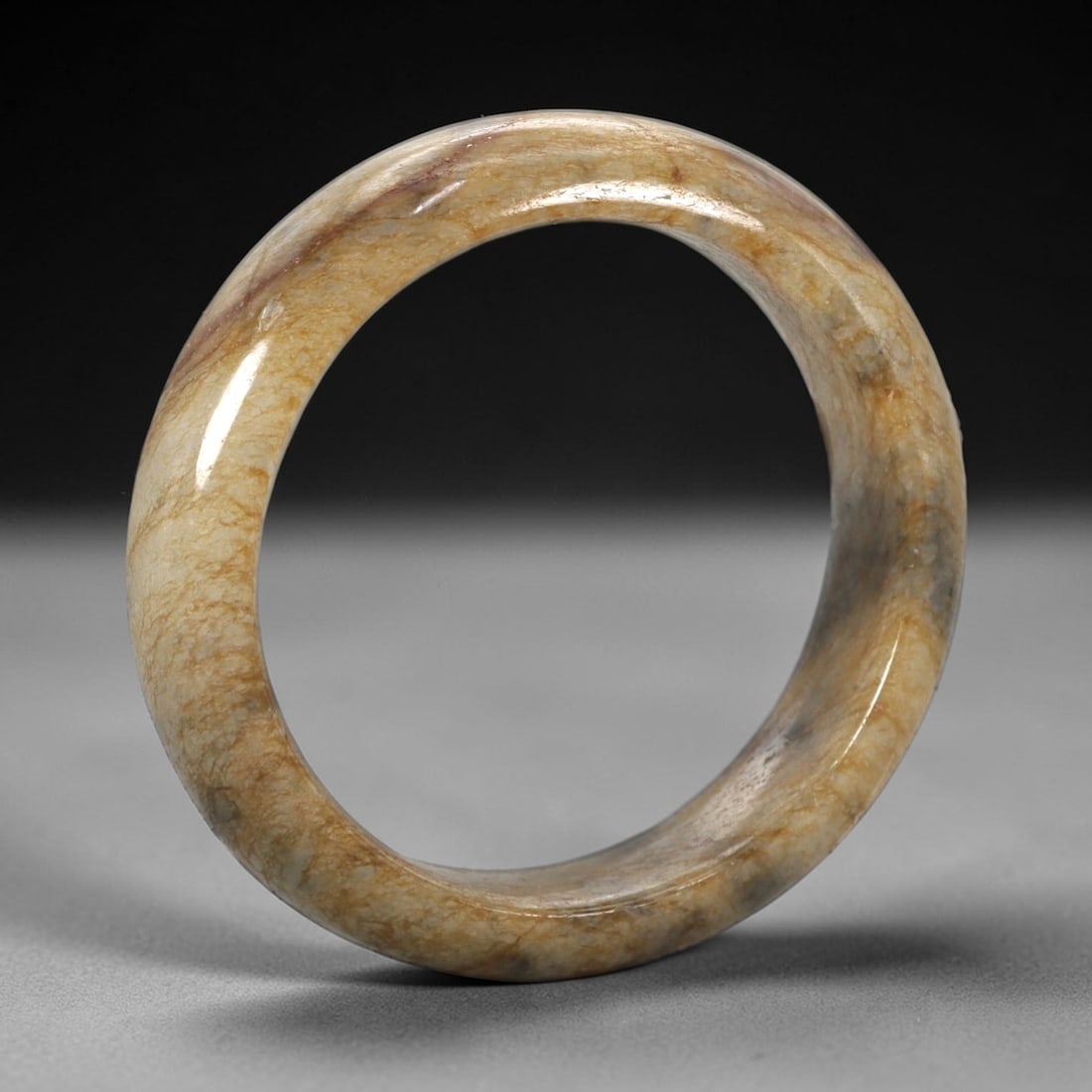 A Chinese Jadeite Jade Bangle Bracelet, 20th Century, 91ZK: Carved from jadeite jade with a pale creamy-tan base and fine, irregular brownish veining throughout, the bangle is formed as a continuous circular band with a rounded cross-section. The surface is sm