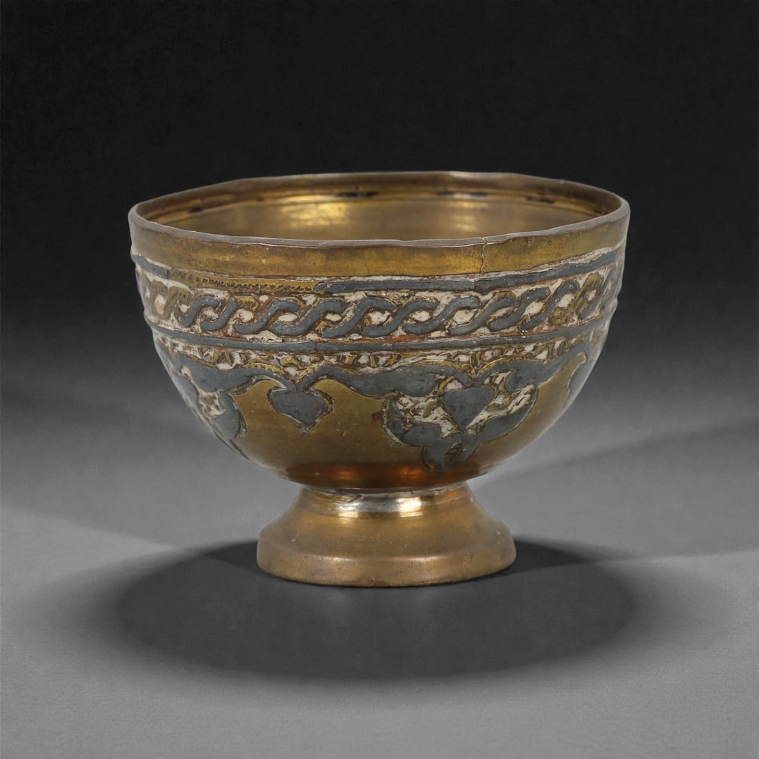 A Brass Bowl, Qajar Period, 19th Century, 58W: Formed from brass and raised on a short circular foot, the bowl has a rounded body with a gently everted rim. The exterior is decorated with engraved and inlaid ornament arranged in horizontal registe