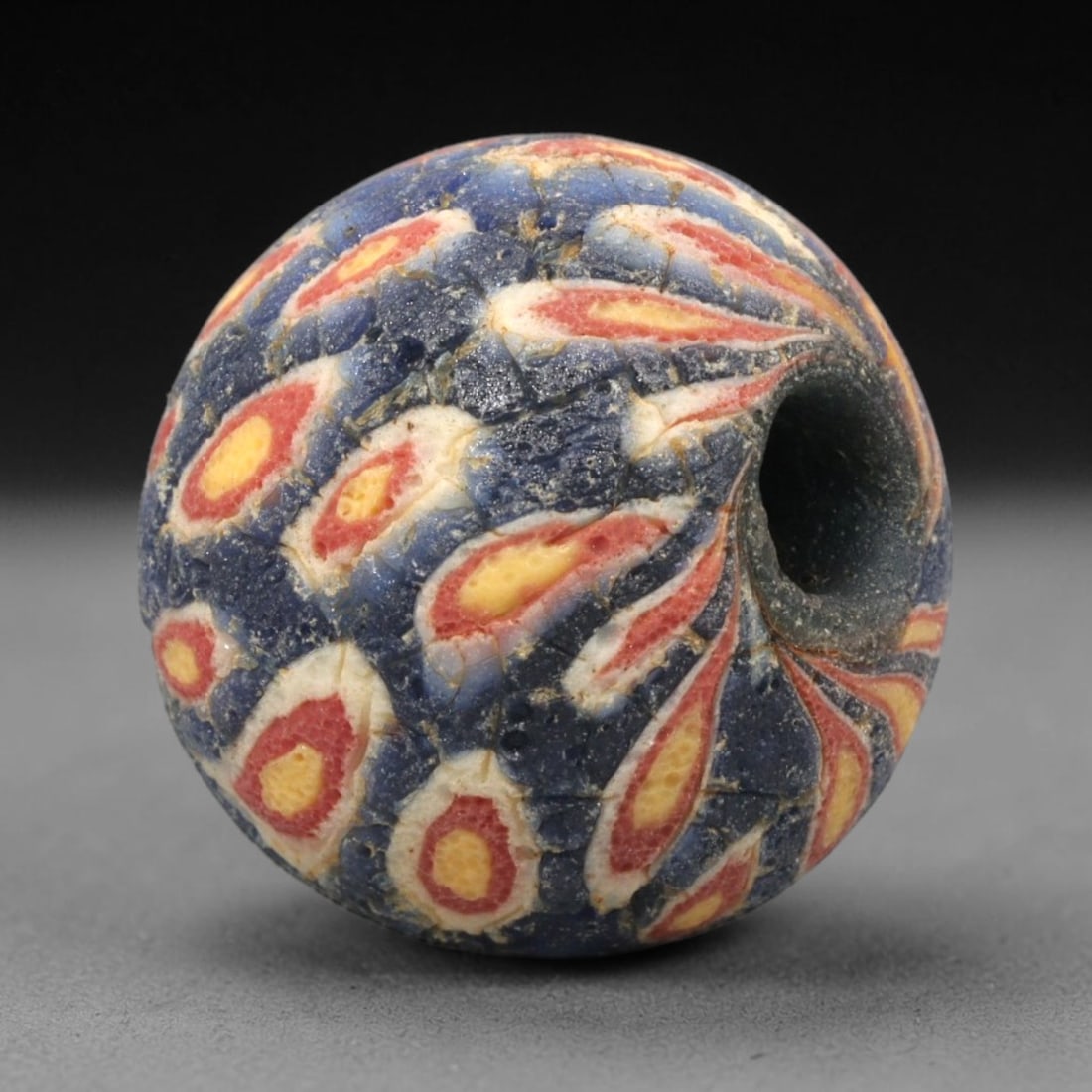 Ancient Mosaic Glass Bead - Roman Peacock Feather Amulet, 45WD: This spherical bead is crafted from core-formed mosaic glass, featuring a deep indigo blue base decorated with repeating peacock feather-inspired motifs. Each motif consists of a central yellow glass