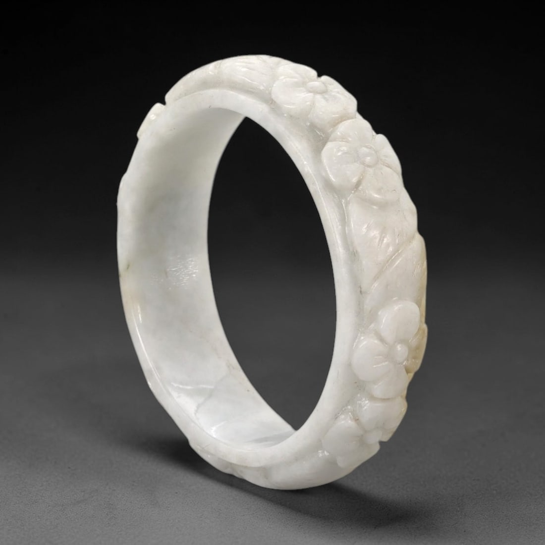 A Chinese Carved Jadeite Jade Bangle Bracelet, 20th Century, 31ZL: A fine jadeite bangle of flattened circular form, masterfully carved in relief with a continuous band of floral blossoms and scrolling foliate motifs. The stone exhibits a translucent pale white base,