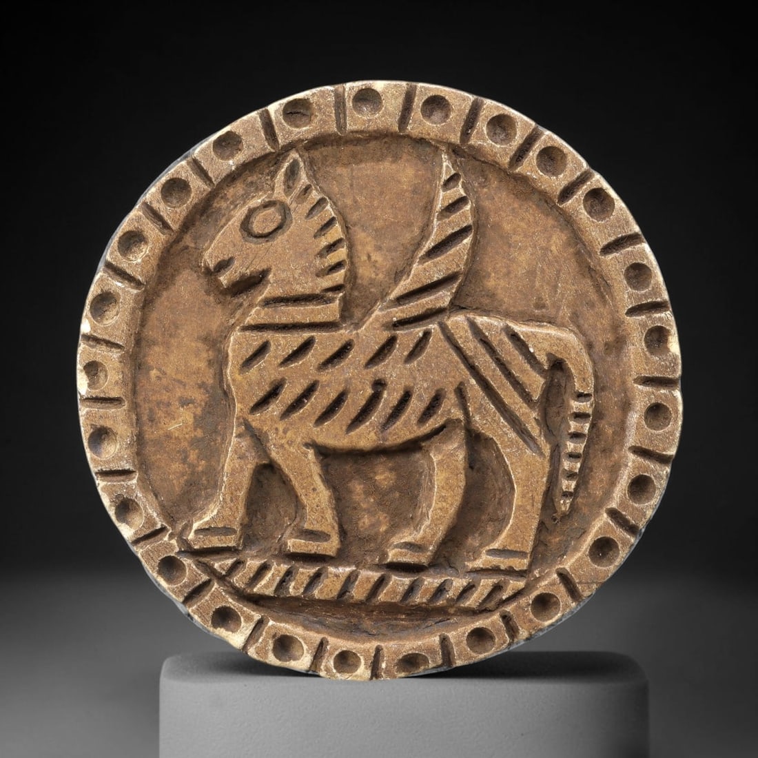 Ancient Steatite Plaque - Near Eastern Winged Animal Relief, 81BE: Carved from steatite, the circular plaque features a prominent raised relief of a winged quadruped, likely a horse or ass, depicted in a striding pose. The creature is rendered with incised linear det