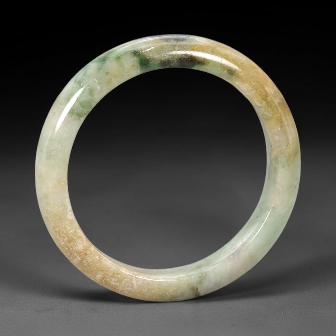 A Chinese Jadeite Jade Bangle Bracelet, 20th Century, 83ZK: Carved from jadeite jade with natural variegated coloration, including pale green, yellowish-brown, and darker green inclusions, the bangle is formed as a continuous circular band with a rounded, slig