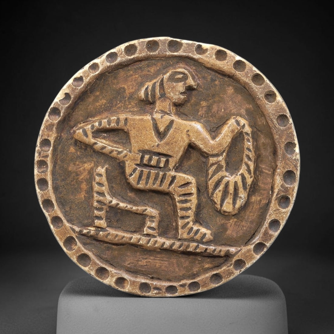Ancient Steatite Plaque - Near Eastern Human Figure Relief, 91BE: Carved from steatite, the circular plaque features a raised relief of a human figure in a dynamic kneeling pose, holding a decorated ring or wreath in one hand. The figure is rendered with incised lin