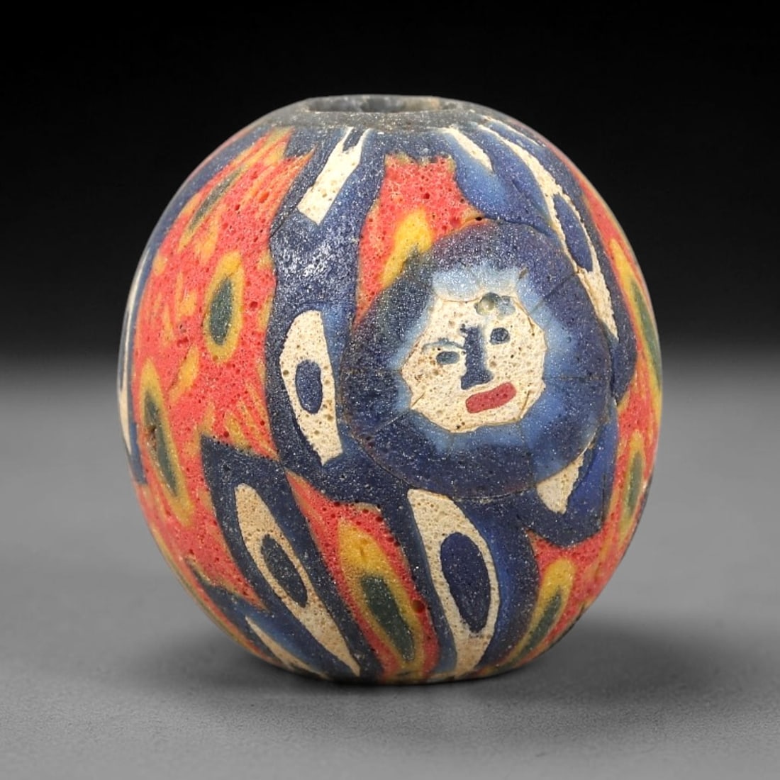 Ancient Glass Bead - Roman Millefiori Face Pendant, 30BG: A spherical ancient glass bead crafted in the millefiori mosaic technique, with a complex multi-layered cane pattern centered on a human face motif. The face is rendered in white glass with blue detai