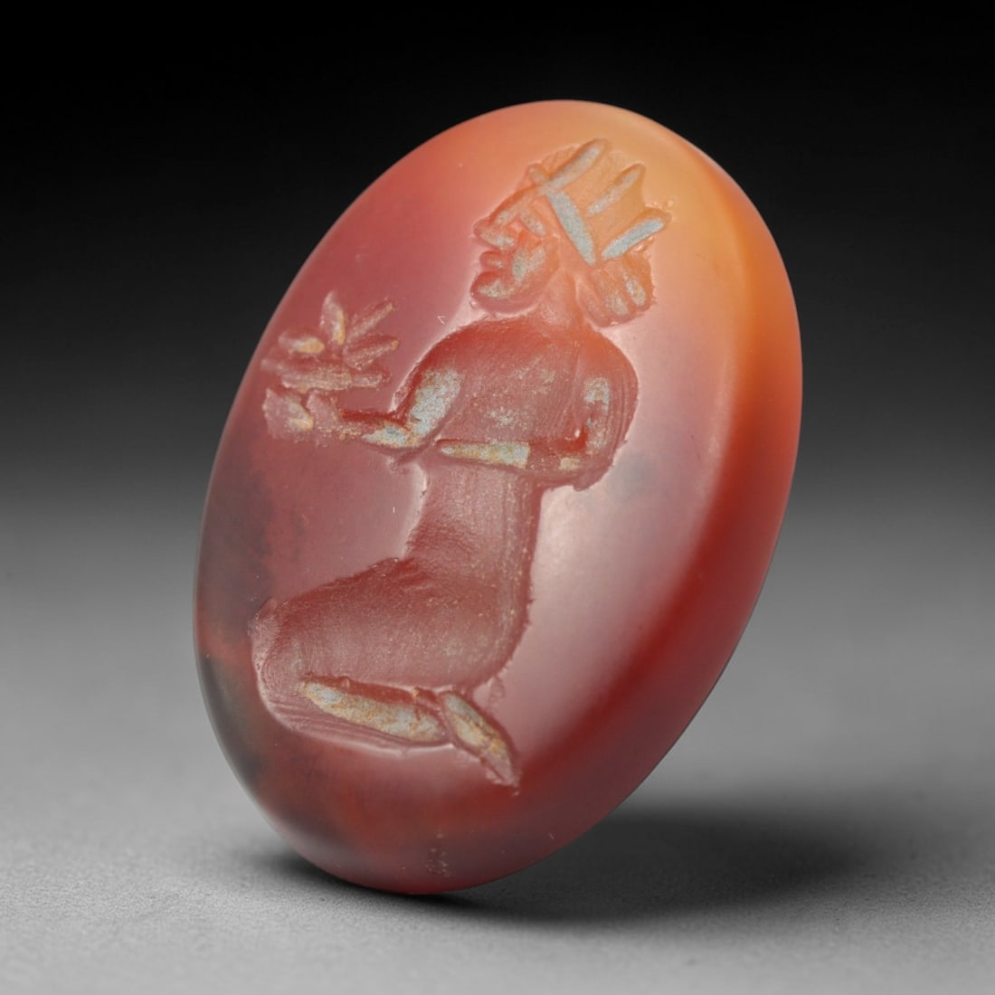 Ancient Agate Intaglio - Near Eastern Kneeling Figural, 40ZW: Carved from warm banded agate, this oval intaglio depicts a kneeling figural form holding a vegetal sprig, adorned with a stylized headdress and incised detailing to denote anatomical structure and de
