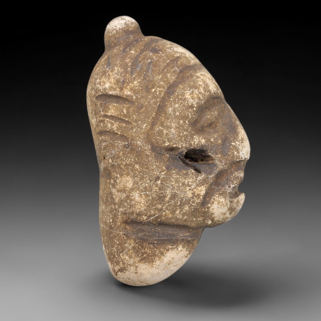 Ancient Stone Bust Amulet - Near Eastern Figurine, 90FX: Carved from a weathered stone, this bust amulet depicts a stylized human head in profile, with incised details defining the facial features, linear head adornments, and a small suspension knob at the