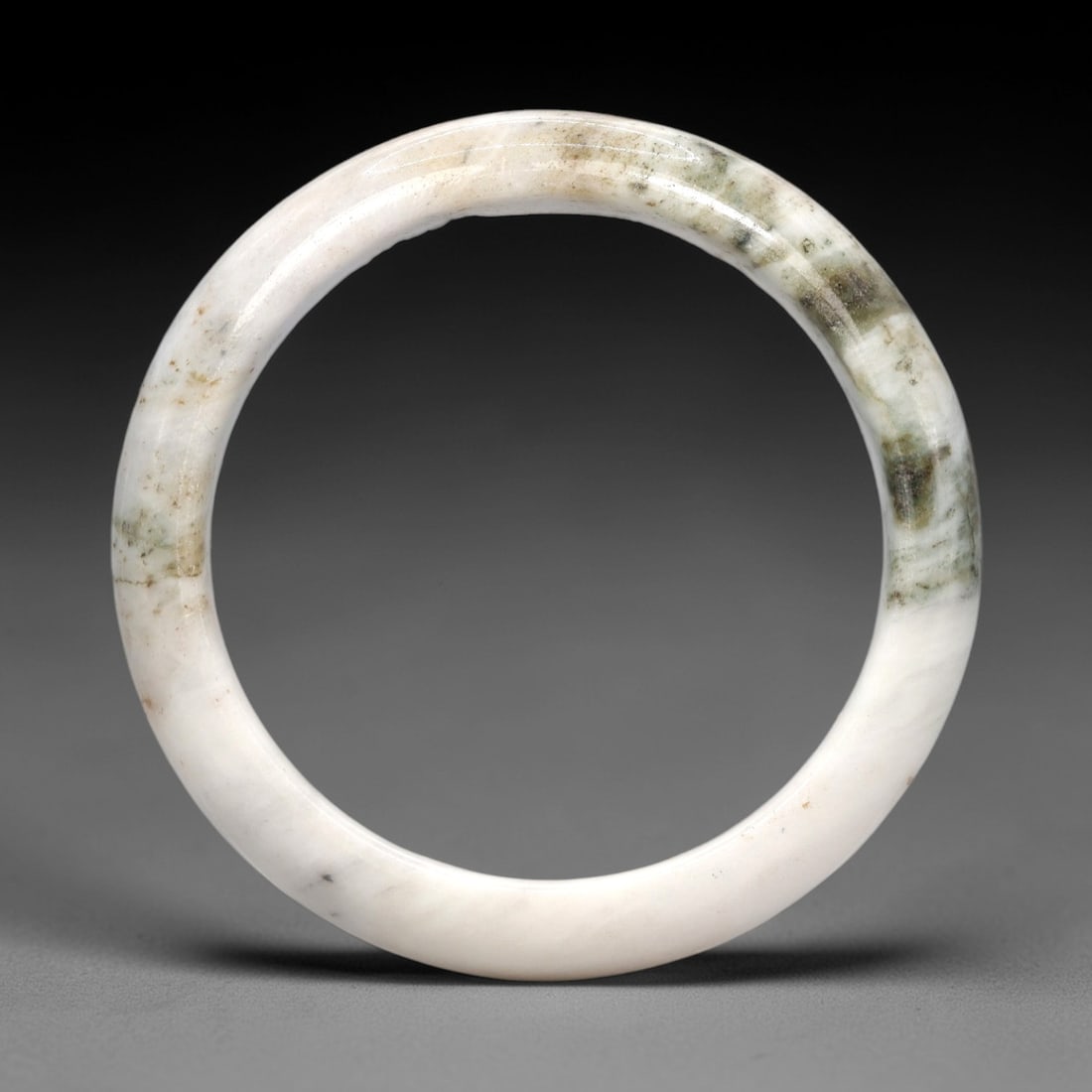 A Burmese Jadeite Jade Bangle, 20th Century, 12FS: A Burmese jadeite jade bangle, with a pale white matrix and prominent dark green mineral inclusions. The bangle is of circular form, with a smooth, polished surface revealing the internal crystalline