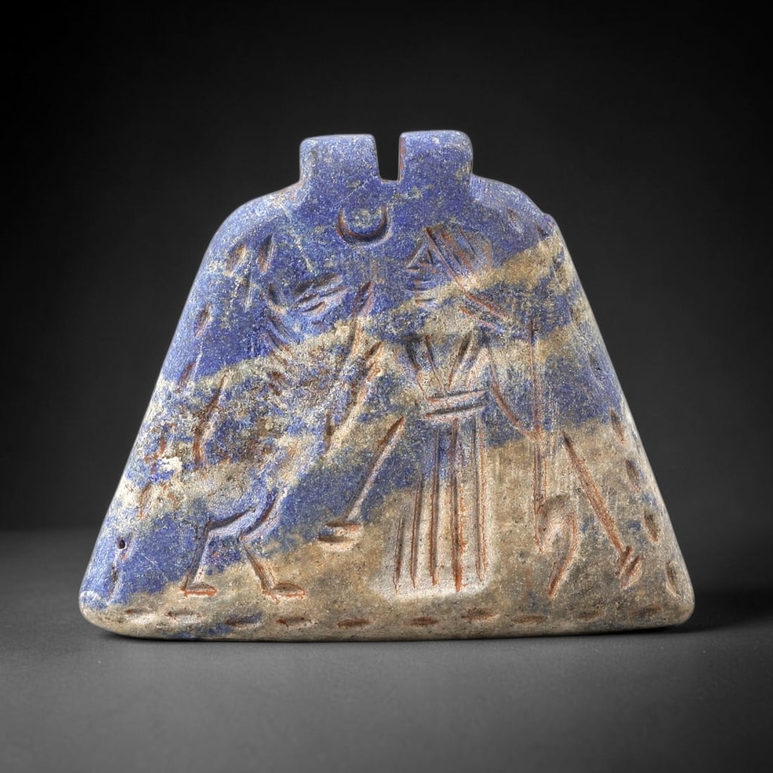 Ancient Lapis Amulet - Near Eastern Figural Scene, 68BG: Carved from lapis lazuli, the amulet is a trapezoidal plaque with two perforated suspension knobs at the upper edge, designed for wearable use. The front face is incised with a figural composition: a