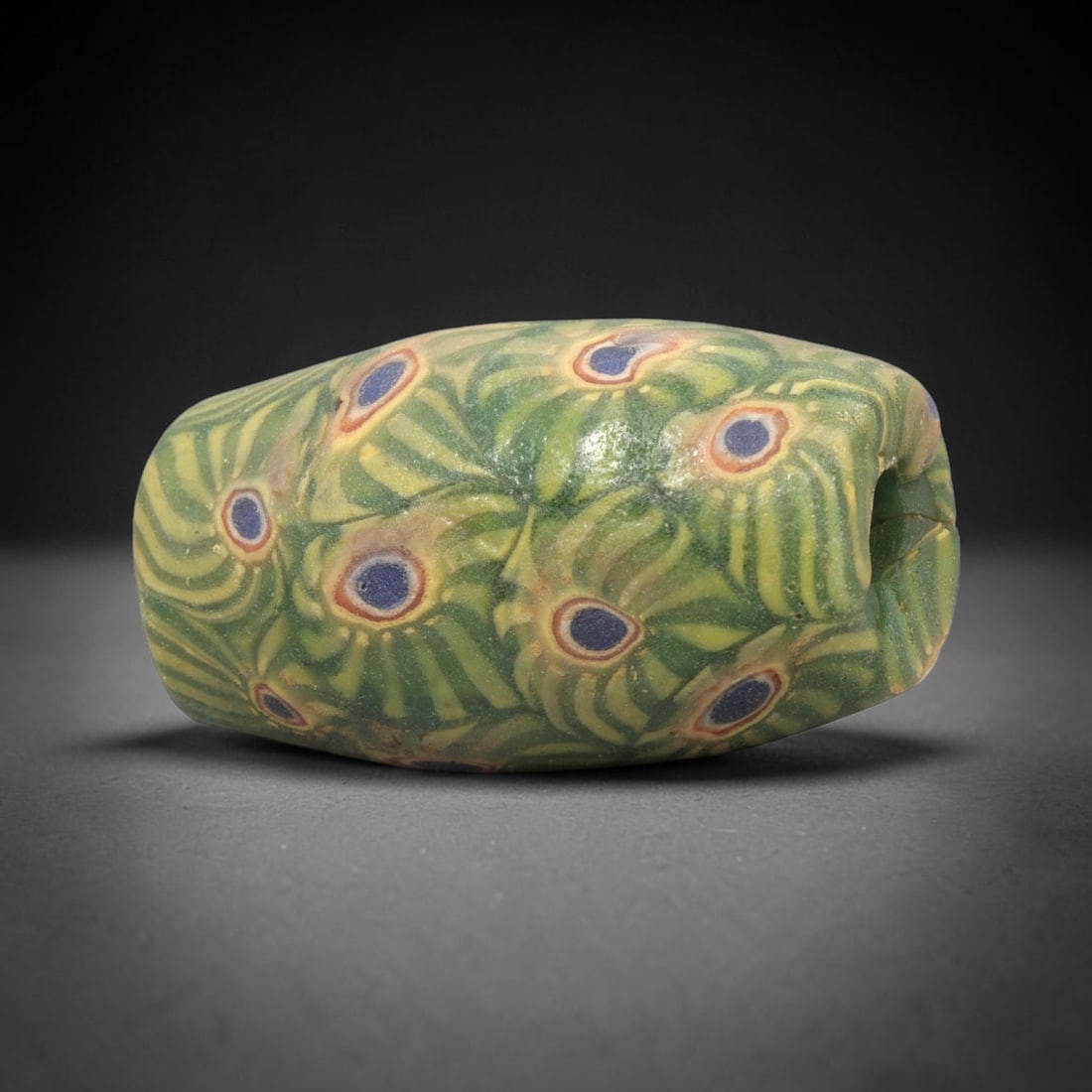Ancient Glass Bead - Roman-Egyptian Millefiori Pendant, 73WD: A cylindrical ancient glass bead crafted in the millefiori technique, with a core of opaque green glass overlaid with intricate cane patterns. The surface is decorated with repeating peacock-eye motif