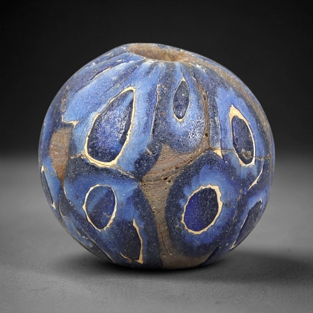 Ancient Glass Bead - Roman Mosaic Spherical Pendant, 31BG: A spherical ancient glass bead crafted using the mosaic glass technique, with a deep cobalt blue glass base adorned with raised teardrop-shaped motifs, each outlined in a thin cream-colored glass laye
