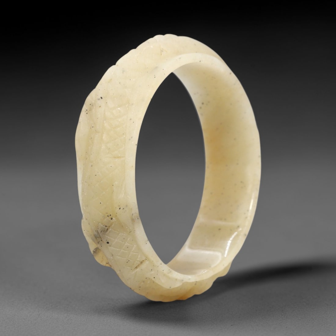 A Chinese Jadeite Jade Bangle Bracelet, 20th Century, 22FS: Carved from pale creamy-yellow jadeite jade with fine dark inclusions, the bangle is formed as a continuous circular band. The exterior surface is decorated with raised relief carving depicting a coil