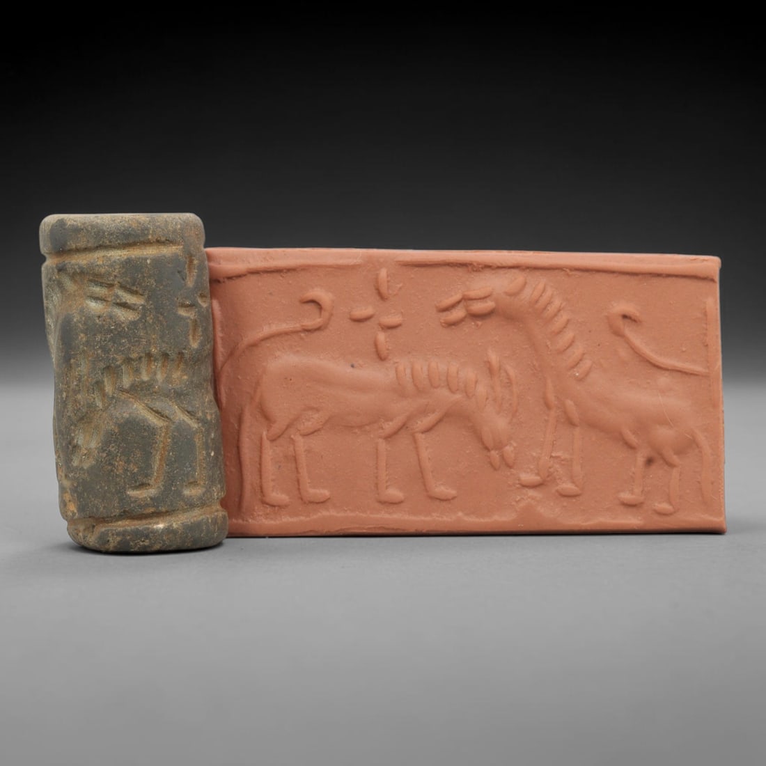 Ancient Stone Cylinder Seal - Mesopotamian Amulet Relic, 58ZA: Carved from a dark, weathered stone, this cylindrical seal features incised decoration depicting two stylized horned quadrupeds in profile, with linear details defining their muscular forms, manes, an
