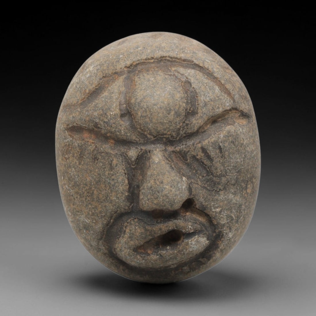 Ancient Stone Maskette - Medieval Human Face Figurine, 34BH: A carved stone maskette of oval form, fashioned from a single piece of weathered grey stone with natural mineral inclusions. The front is carved in deep relief to depict a stylized human face, featuri