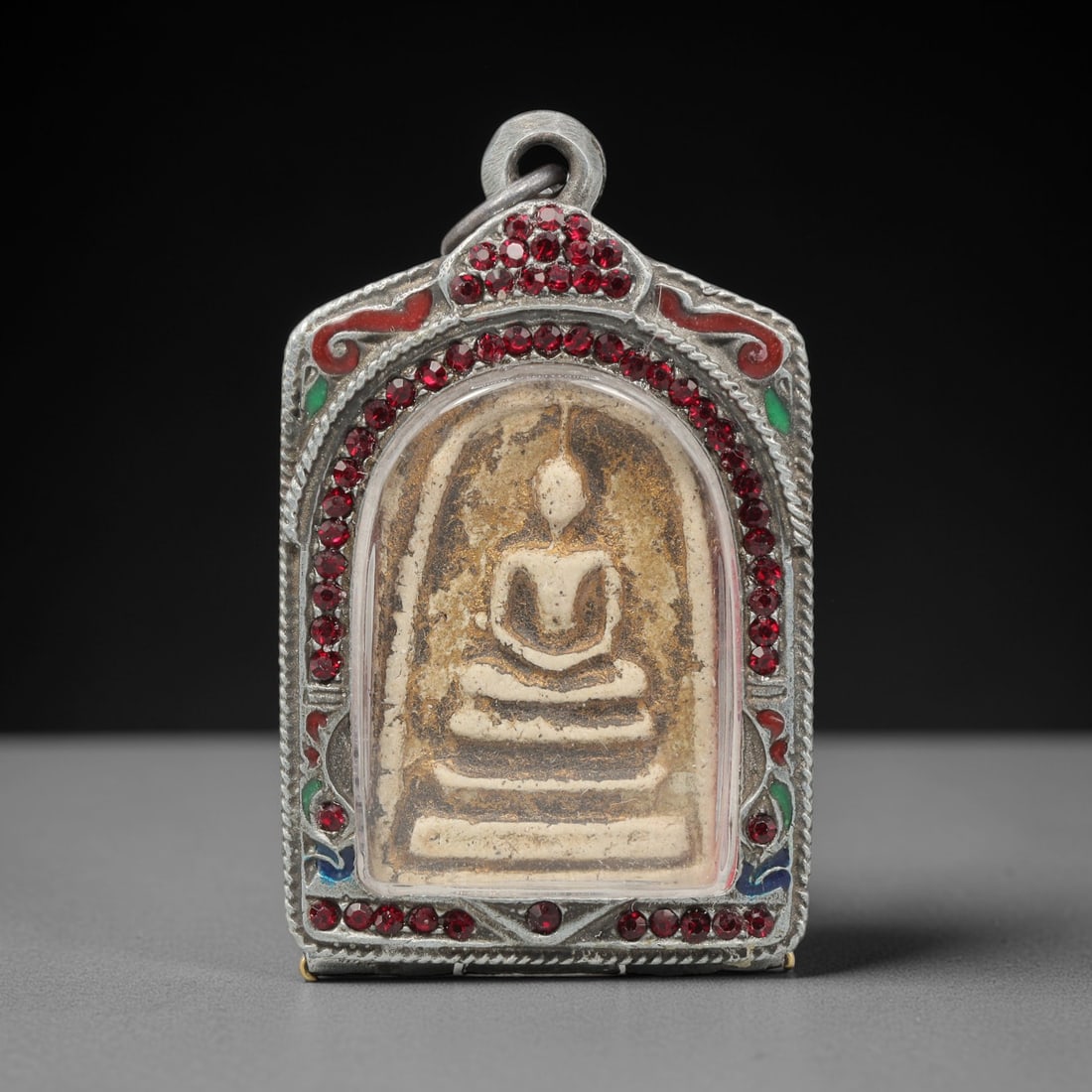 A Thai Terracotta Buddha Amulet in Metal Case, 20th Century, 66ZH: The amulet is formed from terracotta, with a pale beige matrix containing visible particulate inclusions and surface discoloration. The obverse depicts a seated Buddha in meditative posture, seated on