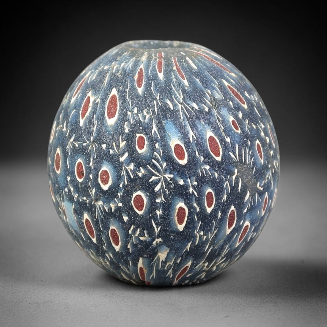 Ancient Glass Bead - Islamic Millefiori Spherical Pendant, 26BG: A spherical ancient glass bead crafted using the millefiori technique, with a deep cobalt blue glass base adorned with intricate cane patterns. The surface features repeating oval motifs, each compose