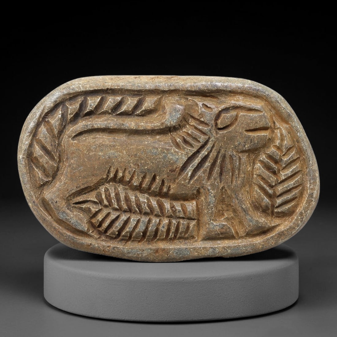 Ancient Stone Plaque Relief - Near Eastern Feline Figurine, 66BE: Carved from a single piece of stone, this oval relief plaque depicts a recumbent feline, rendered in the stylized, linear artistic tradition of the ancient Near East. The figure is detailed with incis