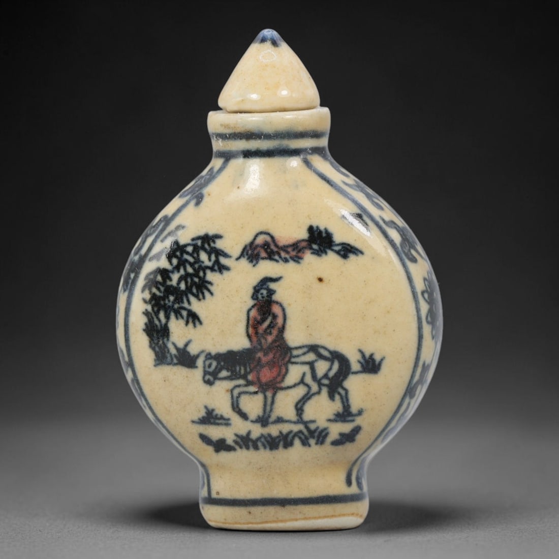 A Chinese Porcelain Snuff Bottle, 20th Century, 7YD: Crafted from porcelain with a pale cream glaze and decorated in underglaze blue with copper red accents, the snuff bottle features a flattened, rounded body, a short cylindrical neck, and a conical st