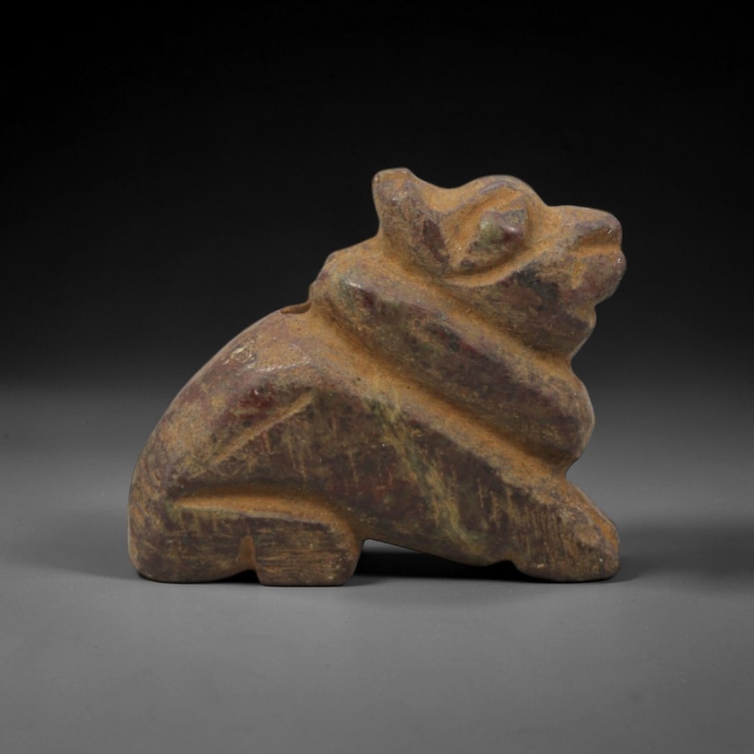 Ancient Stone Feline Amulet - Near Eastern Figurine, 90XV: This amulet is carved from a mottled reddish-green stone with a heavily weathered, earthy patina across its surface, executed in a stylized, angular form that emphasizes the recumbent feline's form. T