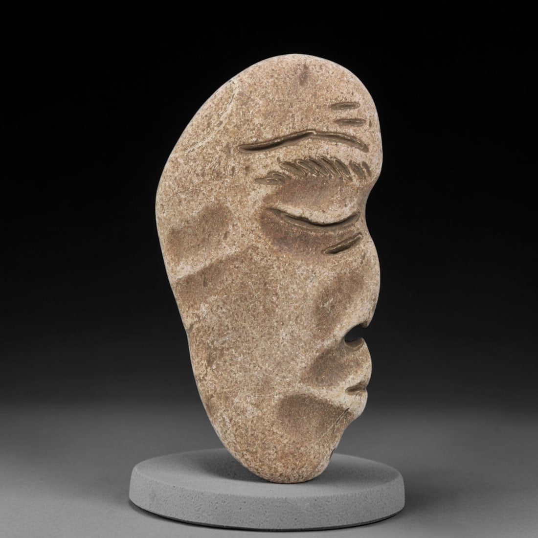 Ancient Stone Maskette - Medieval Human Face Figurine, 53BH: A carved stone maskette of elongated, natural pebble form, fashioned from a single piece of weathered sandstone with a warm tan patina. The front is carved in deep relief to depict a stylized human fa