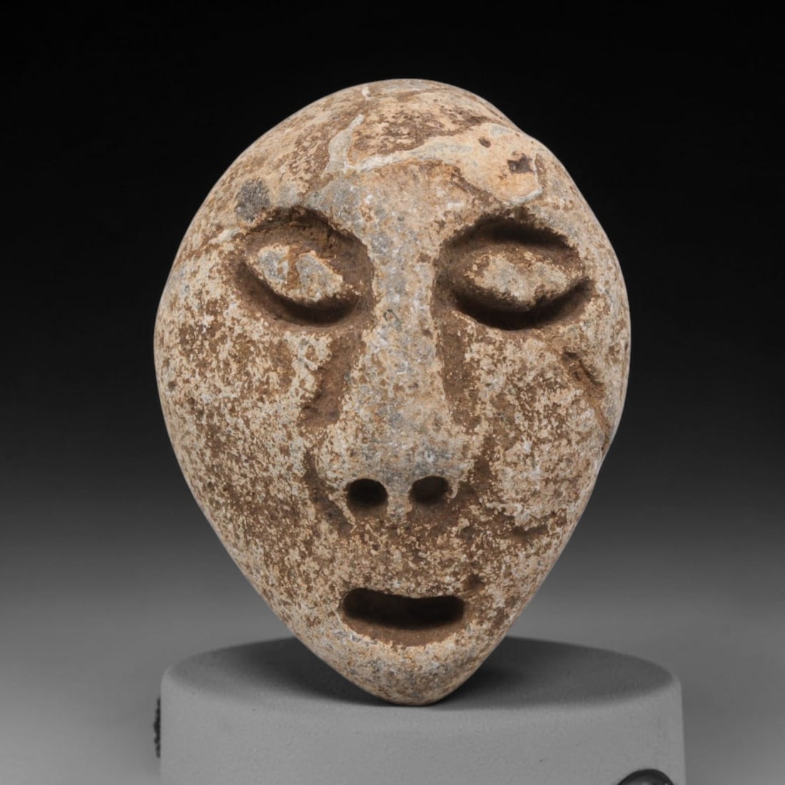 Ancient Stone Maskette - Medieval Human Face Figurine, 40BH: A carved stone maskette of teardrop form, fashioned from a single piece of weathered limestone with natural mineral staining and surface encrustation. The front is carved in deep relief to depict a st
