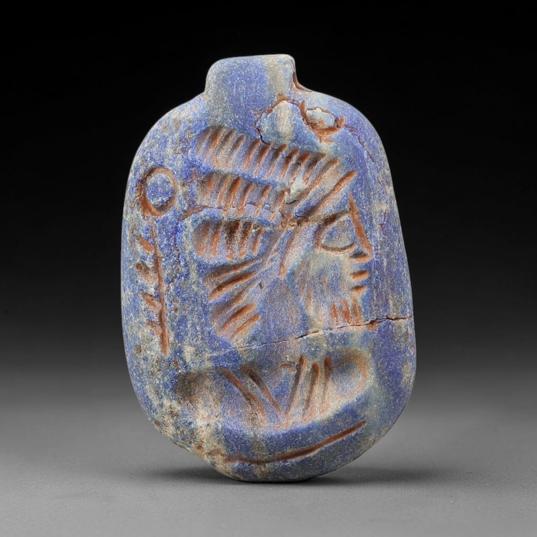 Ancient Lapis Amulet - Near Eastern Figural Pendant, 73BF: Carved from lapis lazuli, the amulet is an oval-shaped plaque with a perforated suspension knob at the upper edge, designed for wearable use. The front face is incised with a stylized human head in pr