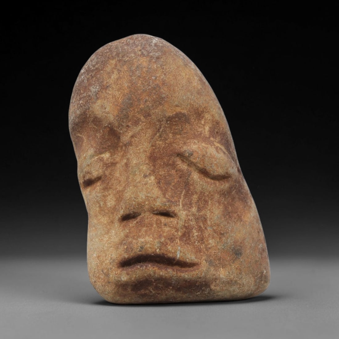 Ancient Stone Maskette - Medieval Human Face Figurine, 41BH: A carved stone maskette of irregular, natural pebble form, fashioned from a single piece of weathered sandstone with a warm tan and brown patina. The front is carved in low relief to depict a serene h