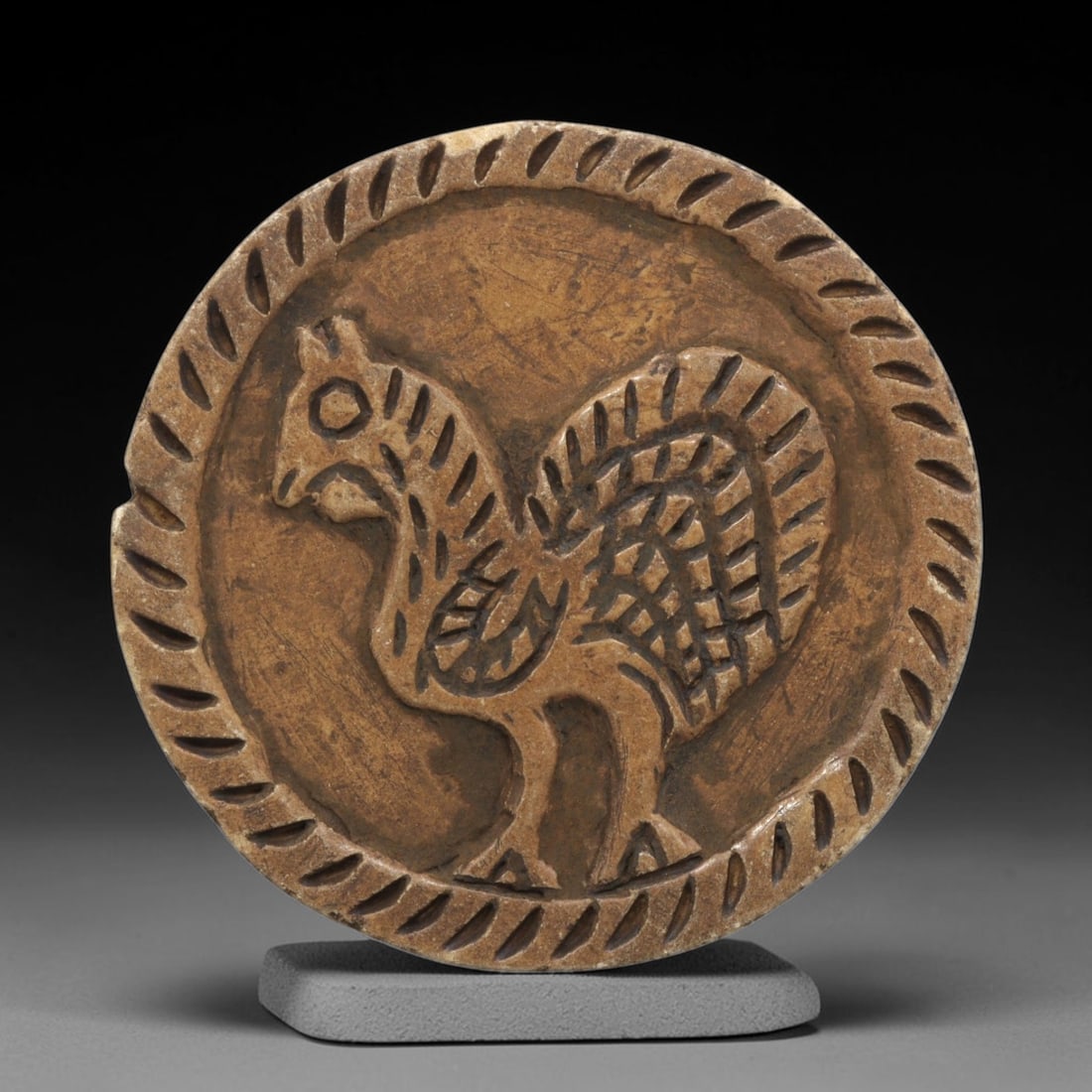 Ancient Steatite Plaque - Near Eastern Bird Relief, 36ZY: Carved from steatite, this circular plaque features a detailed relief depiction of a stylized bird, rendered in the schematic artistic tradition of the Ancient Near East. The figure is shown in profil