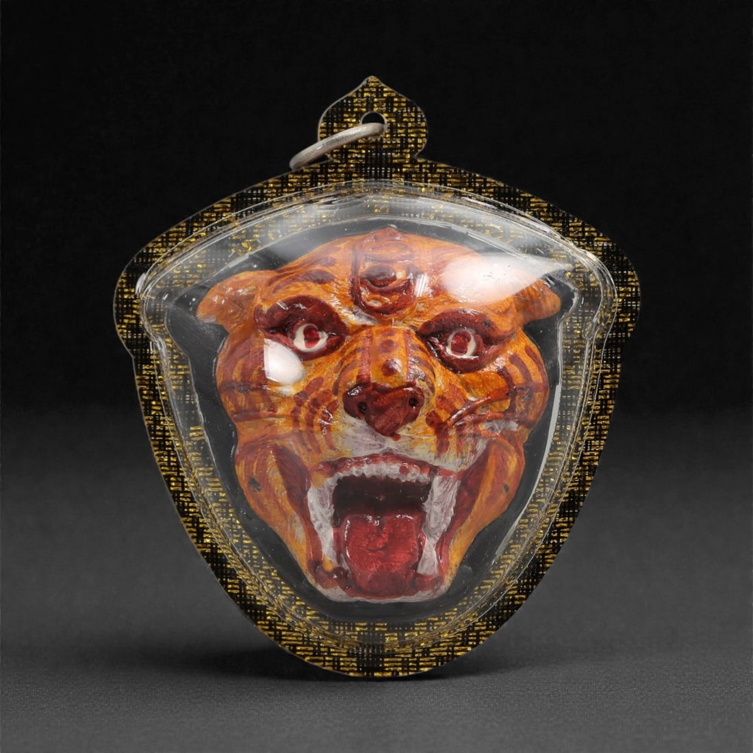 A Thai Painted Tiger Head Amulet in Acrylic Frame, 20th Century, 47FN: The amulet is housed in a shield-shaped acrylic frame with a black-and-gold woven border and a metal suspension loop at the apex. The obverse features a painted high-relief carving of a tiger head wit