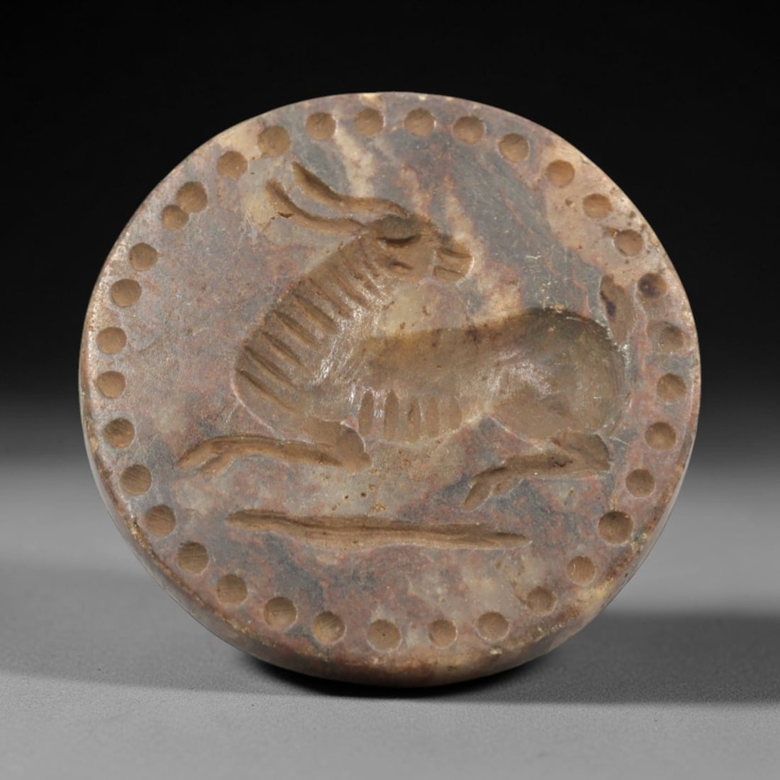 Ancient Near Eastern Stone Ibex Stamp Seal - Amulet, 57ZR: This artifact is a carved stone stamp seal amulet, crafted from a variegated, reddish-brown stone with a smooth, polished surface and natural mineral veining. The circular seal face depicts a recumben