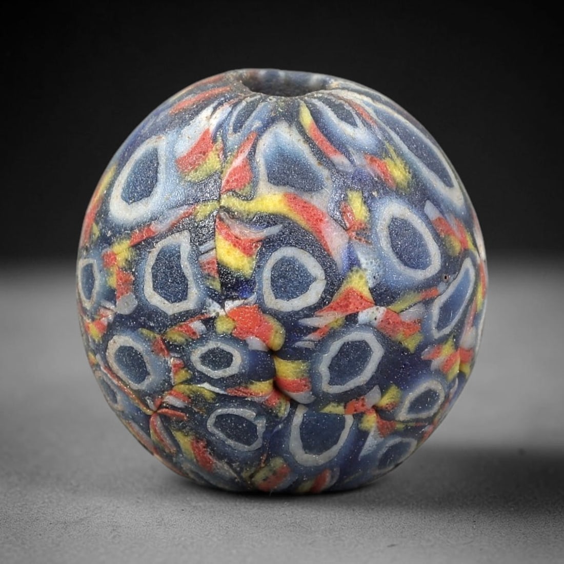 Ancient Glass Bead - Roman Millefiori Spherical Pendant, 36BG: A spherical ancient glass bead crafted in the millefiori mosaic technique, with a deep blue glass base overlaid with intricate fused cane patterns. The surface is decorated with repeating oval motifs,