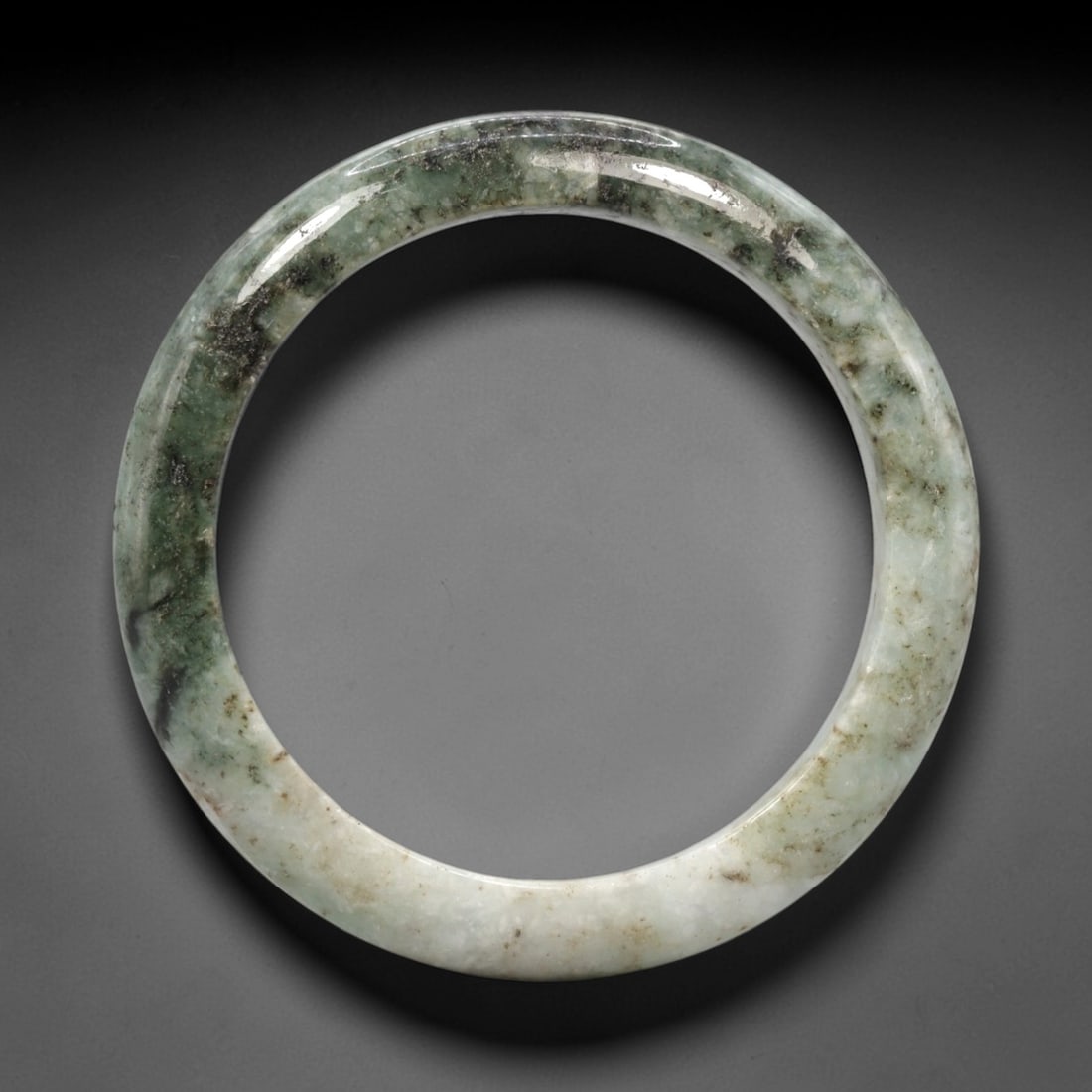 A Chinese Jadeite Jade Bangle Bracelet, 20th Century, 76ZK: Carved from jadeite jade with a pale green base and a distinct dark green section at the upper portion, the bangle is formed as a continuous circular band with a rounded cross-section. The surface is