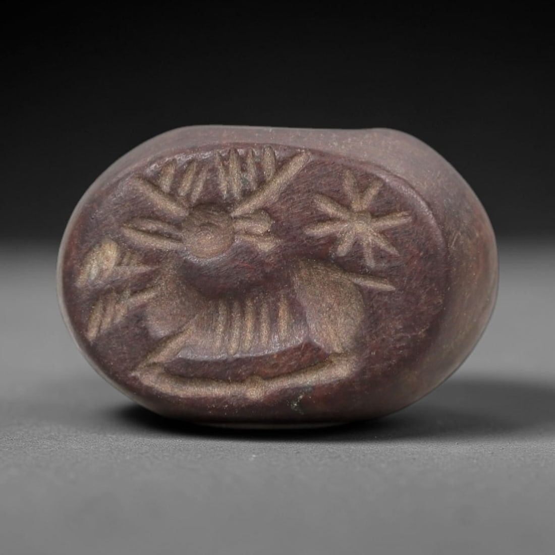 Ancient Stone Hemispherical Stamp Seal - Near Eastern Stag Figure, 17AN: Carved from a dense, dark reddish-brown stone, this hemispherical stamp seal features a flat, oval face incised with a stylized stag in profile, accompanied by a radiate star motif. The stag is render